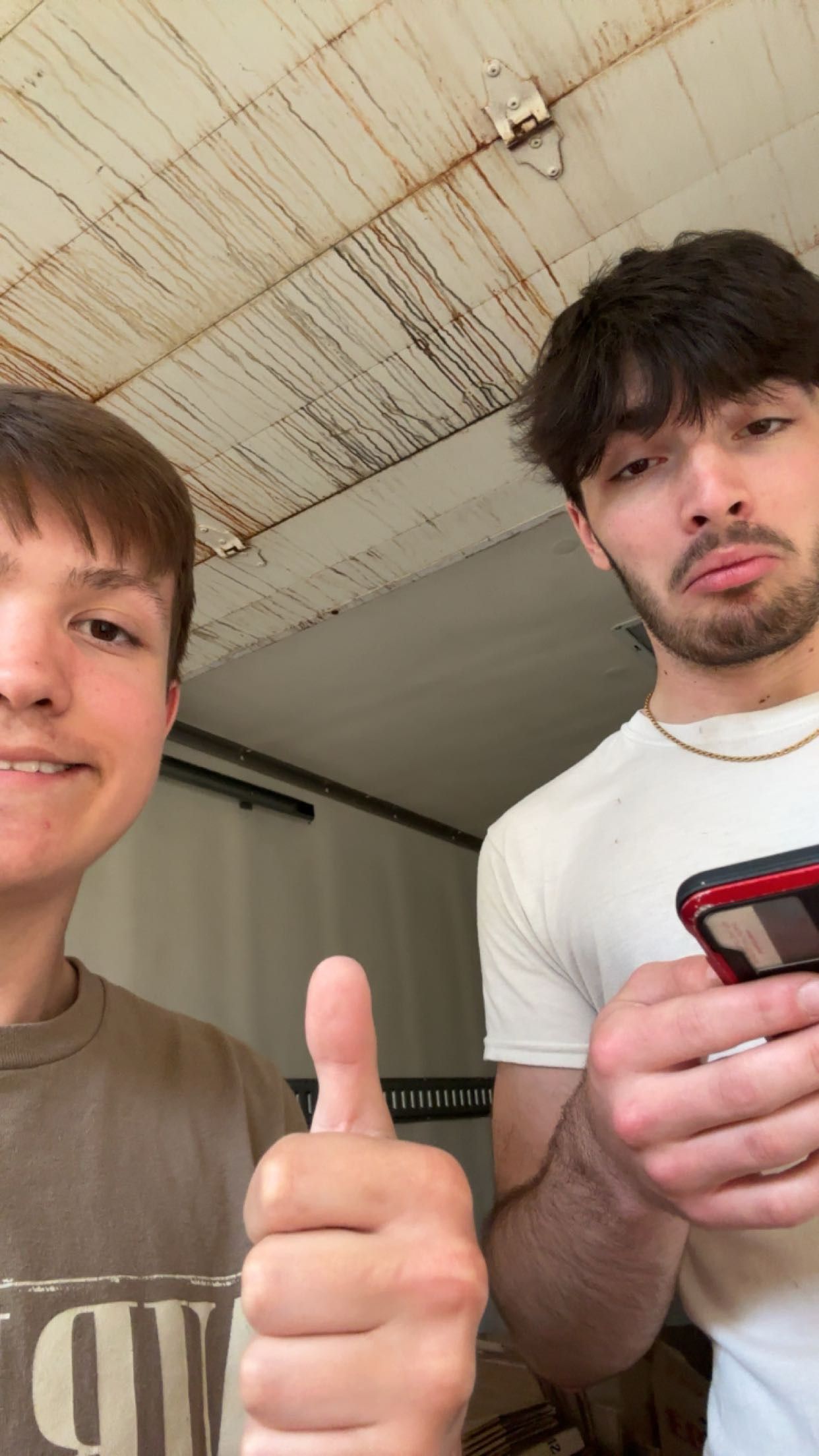 Two young men taking a selfie indoors, one giving thumbs up, the other holding a red phone, wearing casual t-shirts.