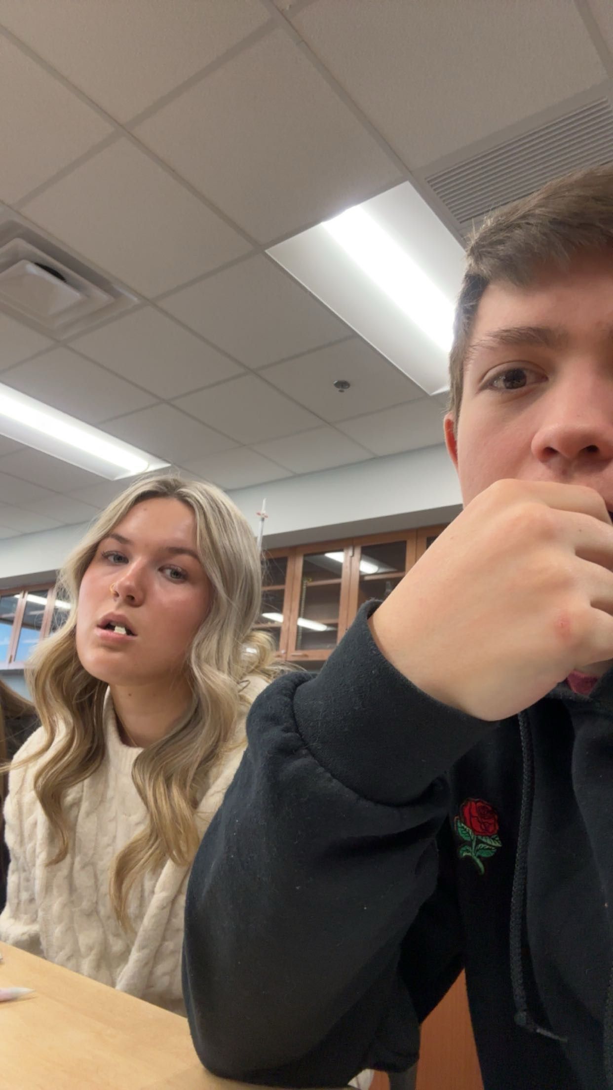 Two students sitting at a desk in a classroom, a blonde girl in a white sweater and a boy in a dark rose-logo hoodie.