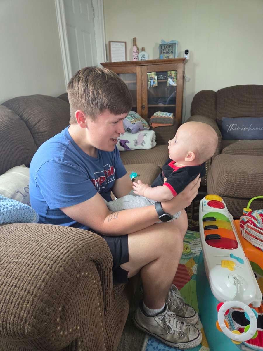 Young man sitting on couch holding and interacting with a smiling baby in a cozy living room setting.