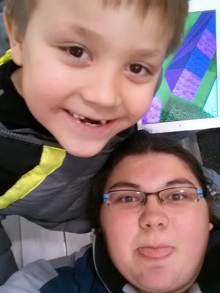 A young smiling boy with a missing tooth takes a selfie with a woman wearing glasses indoors.