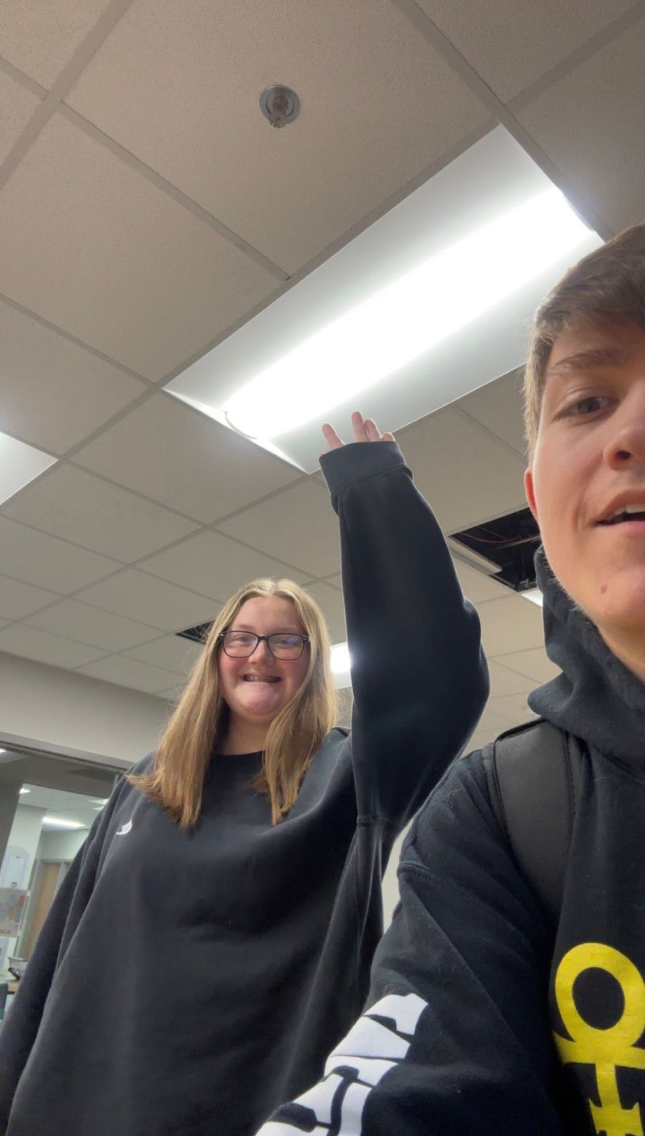 Two teens take a selfie in a classroom, one raising their hand and making a funny face.