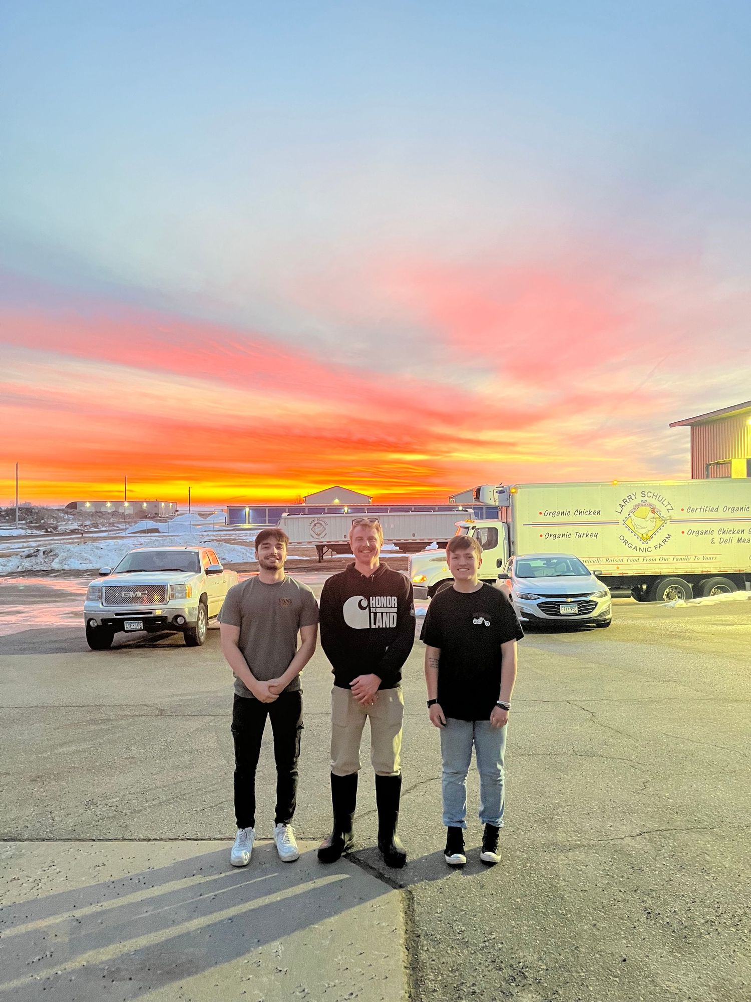 Three people posing together outdoors at sunset with vibrant orange and pink sky, vehicles and a truck visible behind them.