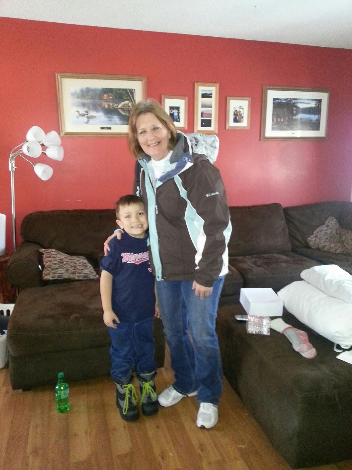 A young boy in a Twins baseball uniform poses with an older woman in a brown jacket in a red-walled living room.