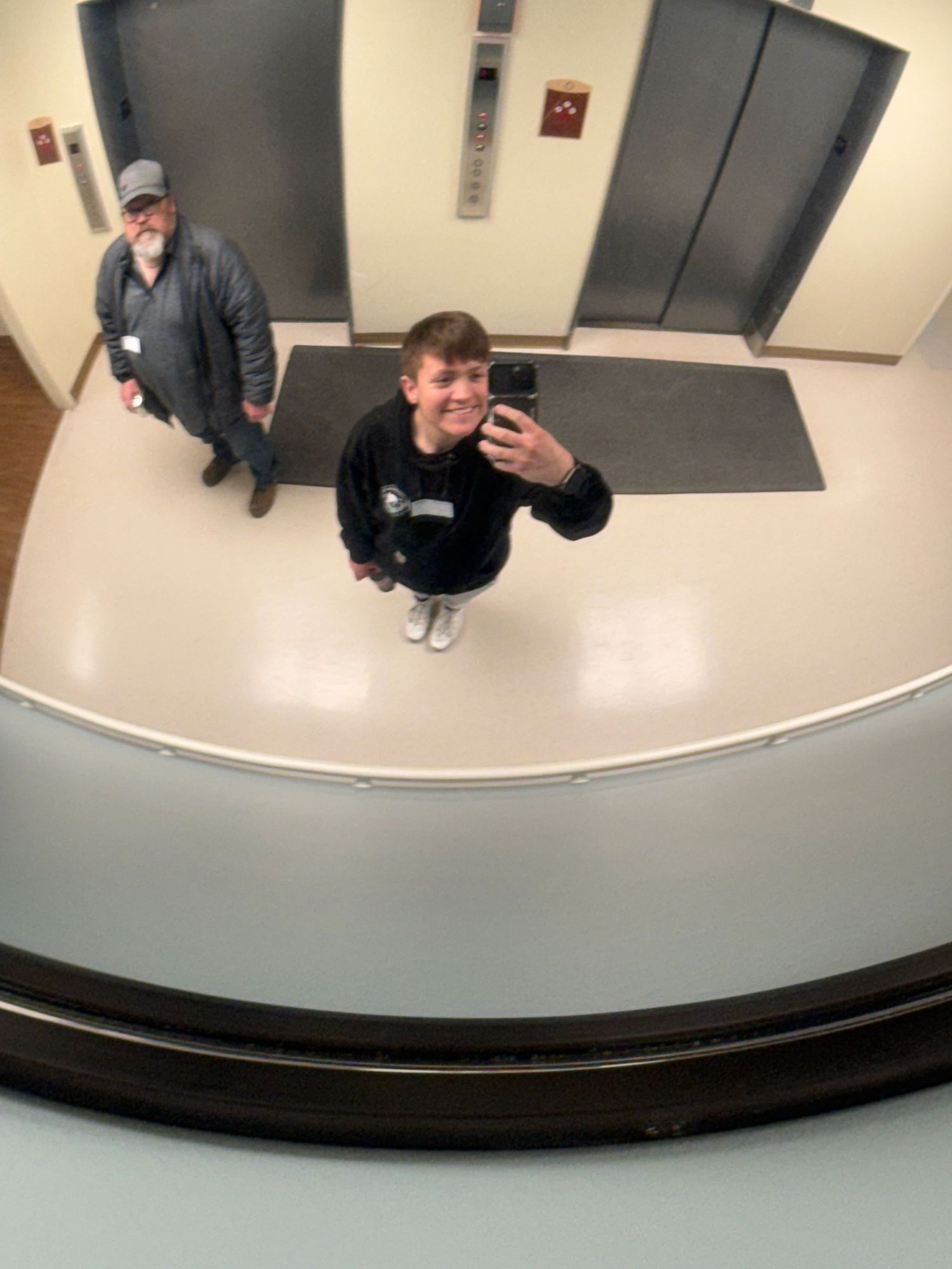 Two people taking a mirror selfie in an elevator lobby, one smiling at camera holding phone, another standing in background.
