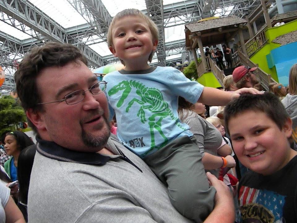 A man holds two young boys on his shoulders at a busy indoor event, smiling for the camera.