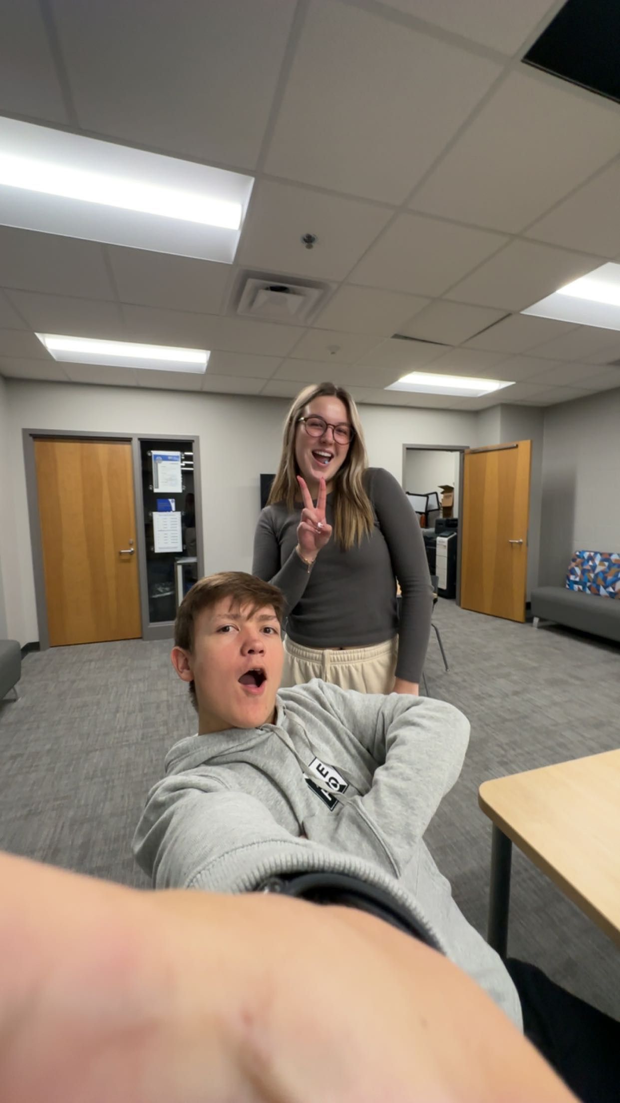 A young man in a gray hoodie makes a funny face while a smiling woman flashes a peace sign in an office setting.