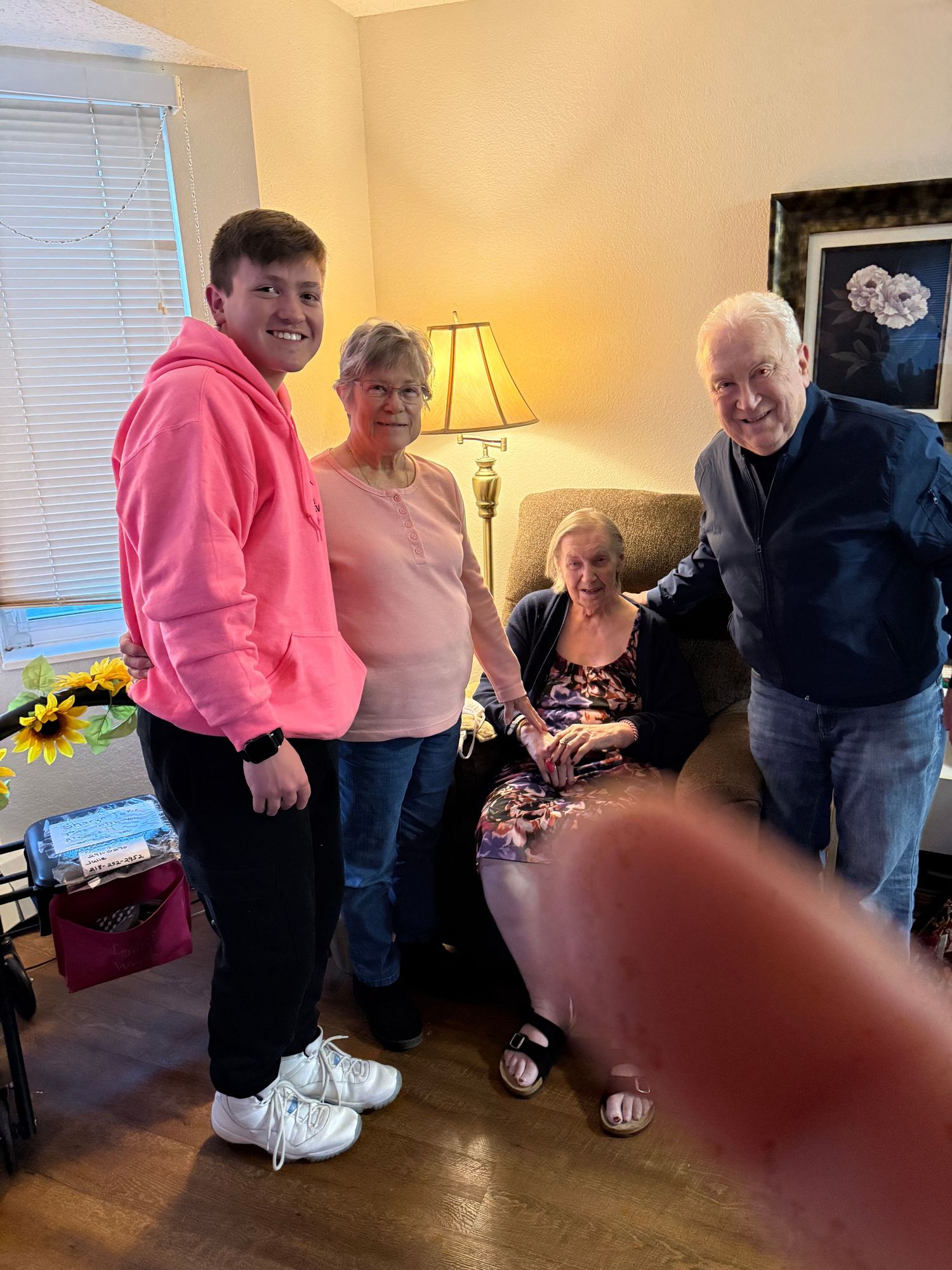 Four people posing together in a cozy living room, one wearing a pink hoodie, gathered around a seated elderly woman.