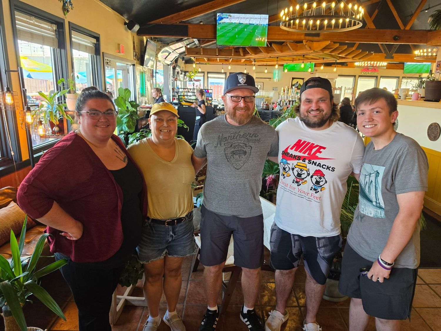 Five friends pose together smiling inside a casual restaurant with green plants and a TV in the background.