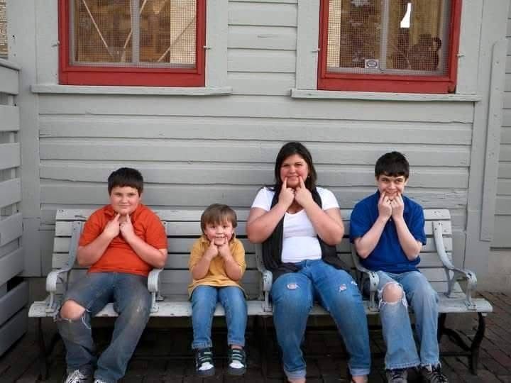 Four children sitting on a bench outside a gray building making funny faces.
