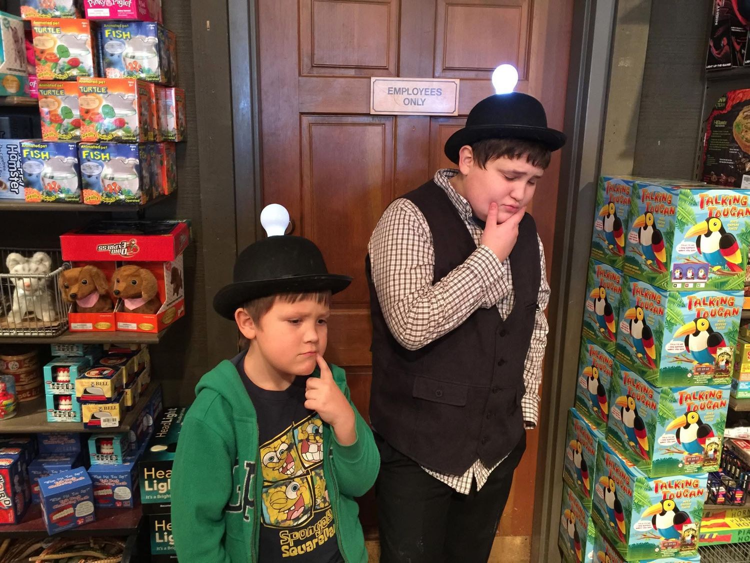 Two boys wearing mining hats pose thoughtfully in a colorful toy and game store near an employees only door.