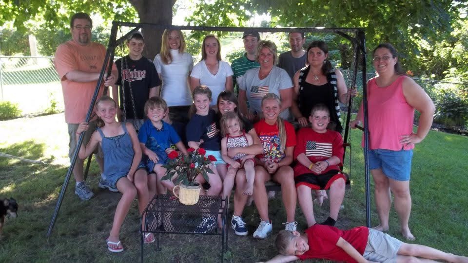 Large family group photo outdoors on a sunny day, with adults and children posing together near a swing set on green grass.