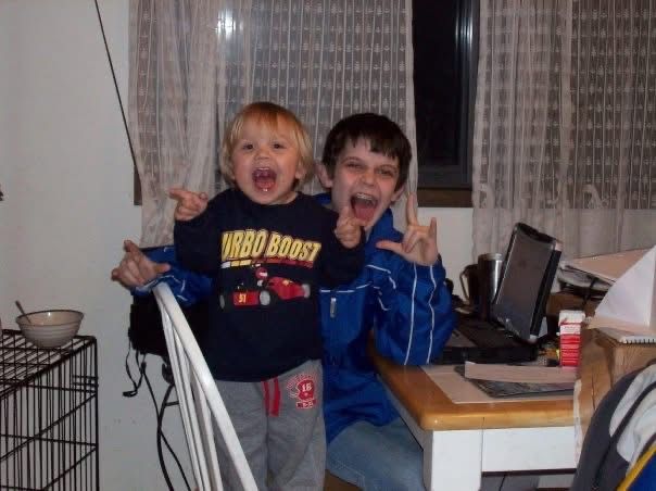 Two young boys making rock on hand gestures and silly faces while standing near a desk with a laptop.