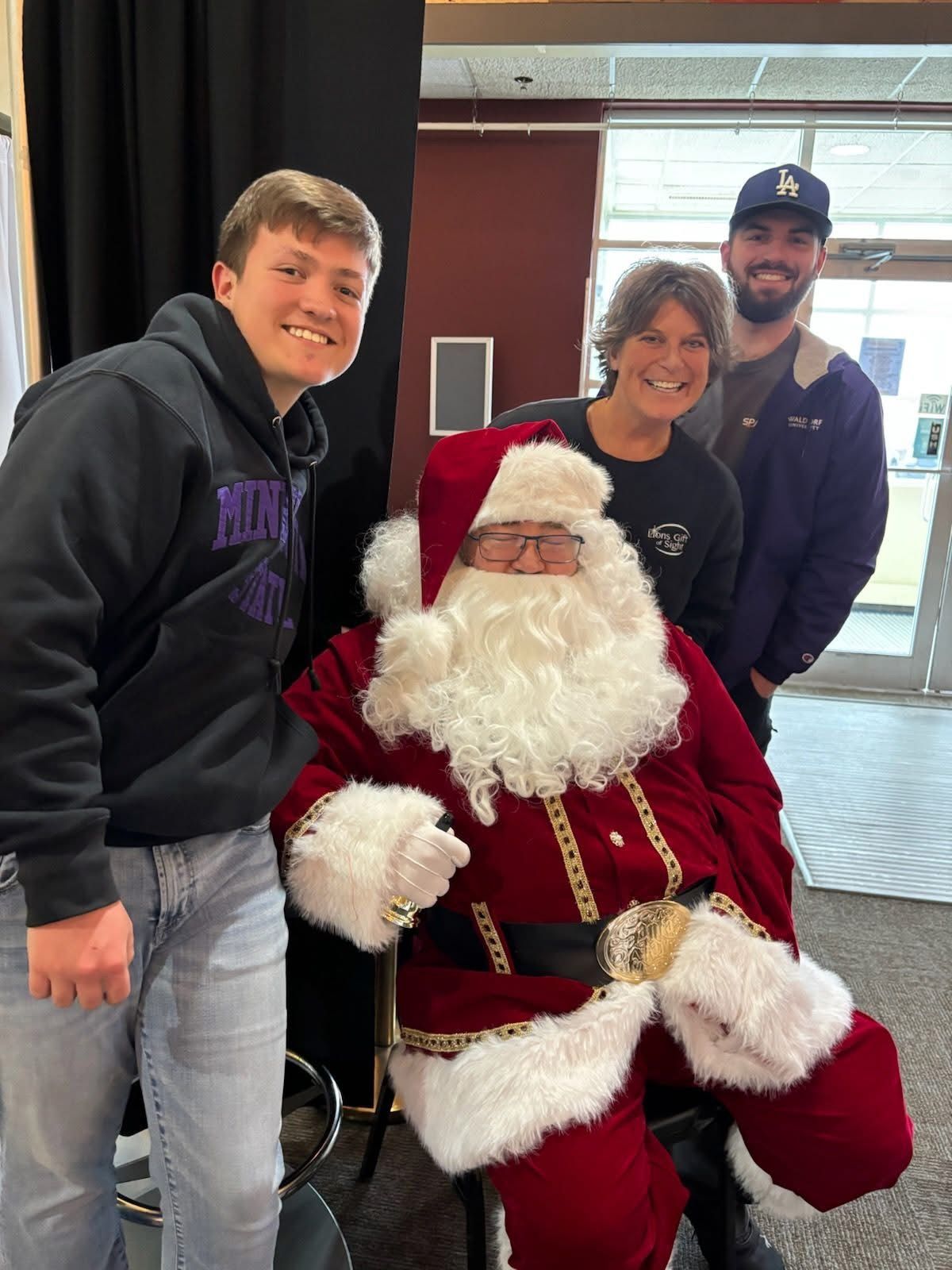 Three people pose with Santa Claus seated in a red suit at a festive indoor holiday event.