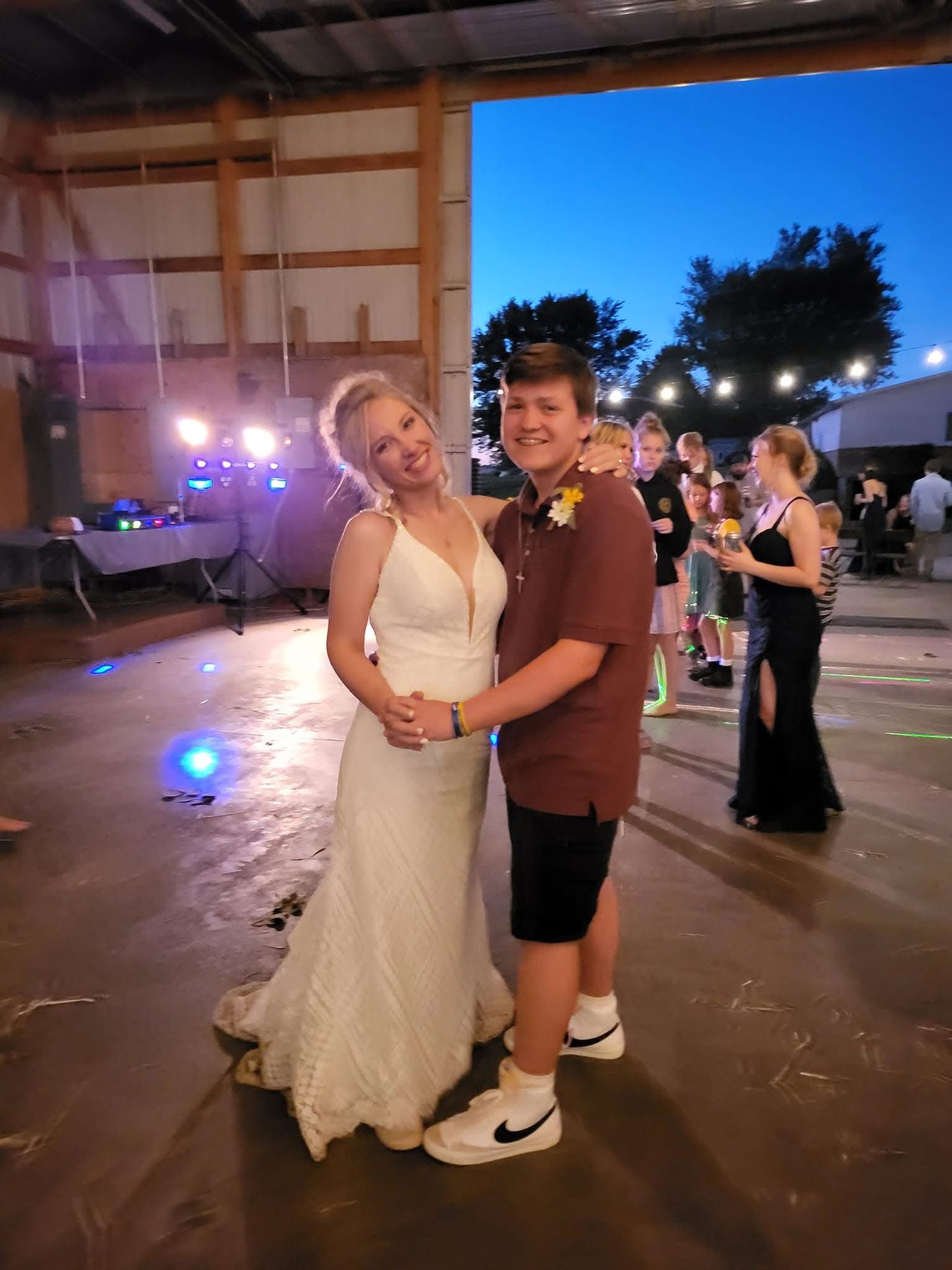 Bride in white gown poses with young guest on dance floor at rustic barn wedding reception at dusk.