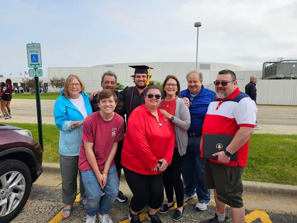 Family group photo celebrating a graduation outdoors, with graduate in cap and gown surrounded by smiling relatives.