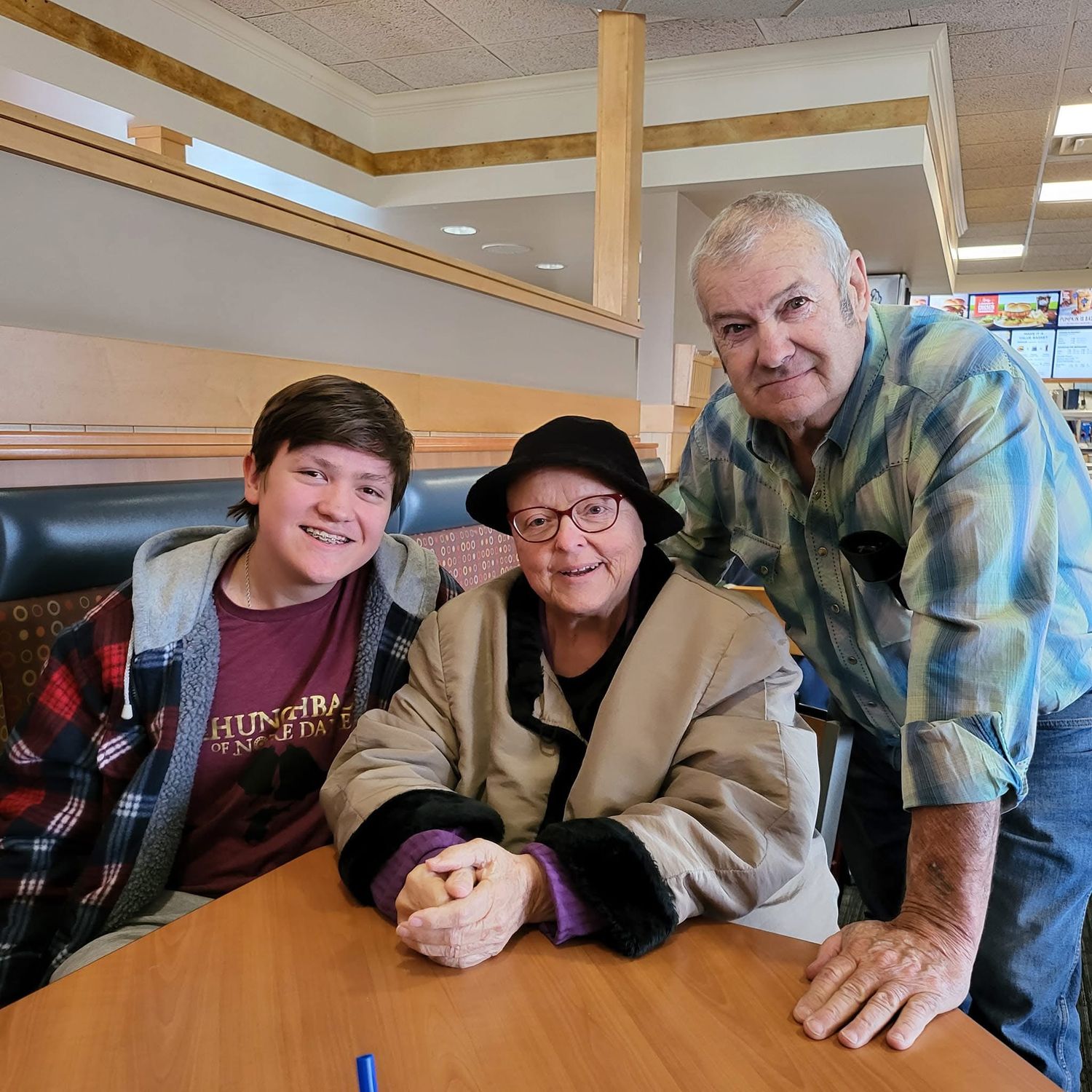 Three generations of family smiling together at a restaurant table, including a young teen, elderly woman in hat, and older man.