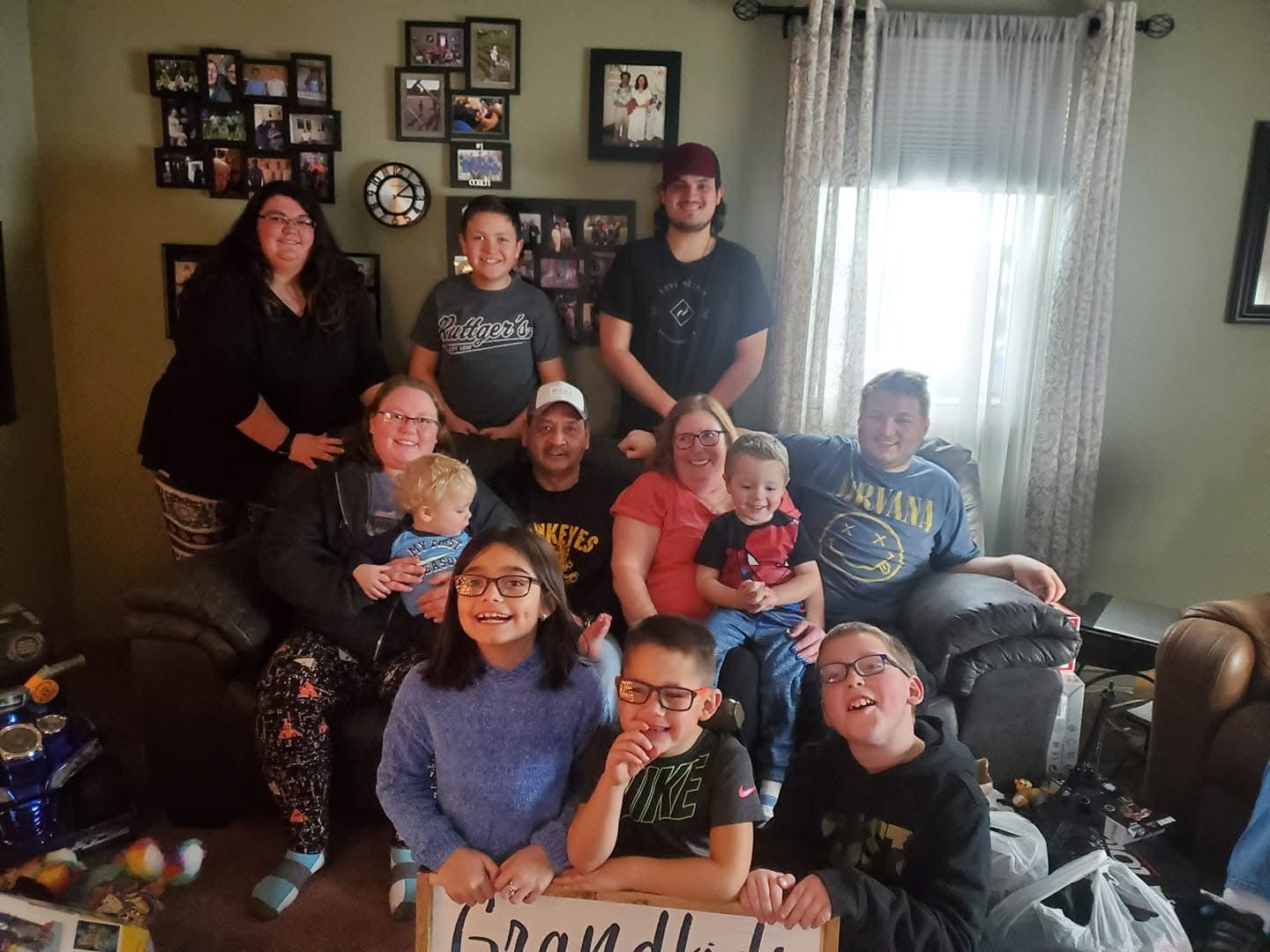 Large family group photo indoors with grandchildren holding a sign, multiple generations smiling together in a living room.
