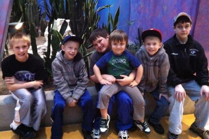 Six young boys sitting together on a bench, smiling for a group photo indoors with tropical plant decor.