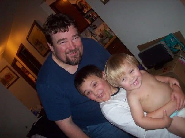 Man smiling with two young boys hugging in a warmly lit home setting.