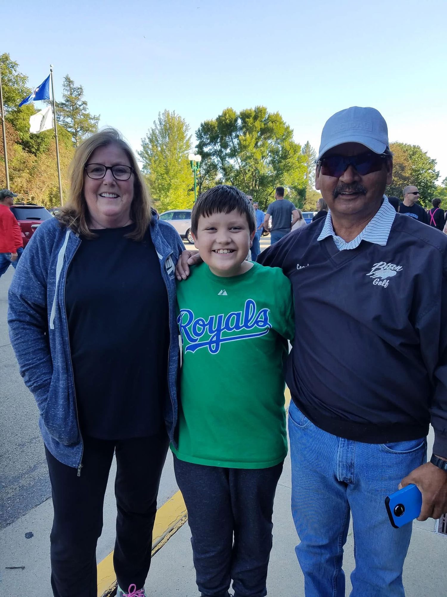 A woman, young boy in Royals shirt, and older man in cap smile together outdoors at a sunny event.