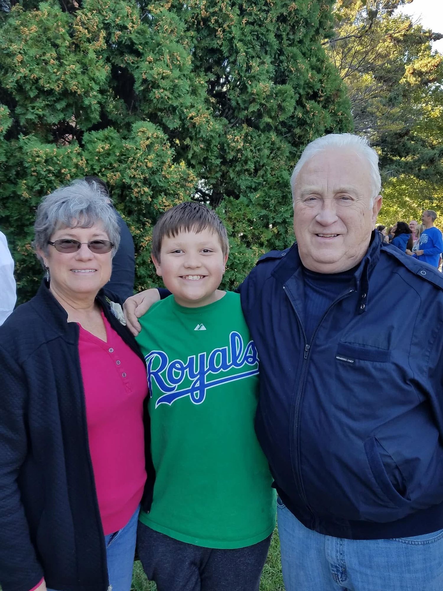 A boy in a Kansas City Royals shirt smiles between two adults outdoors near green trees.