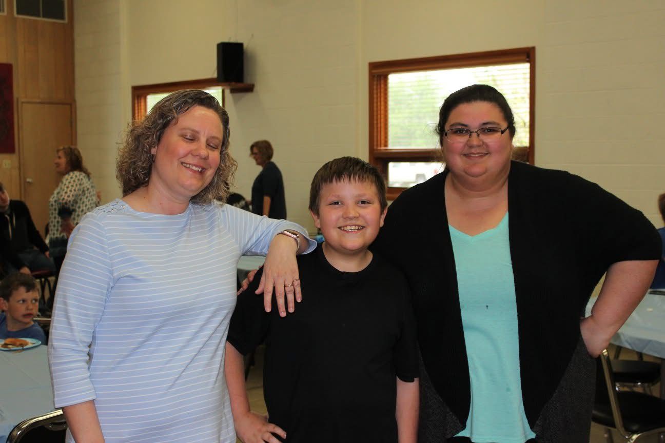Two women and a young boy smile together at an indoor gathering, standing near tables in a community hall.