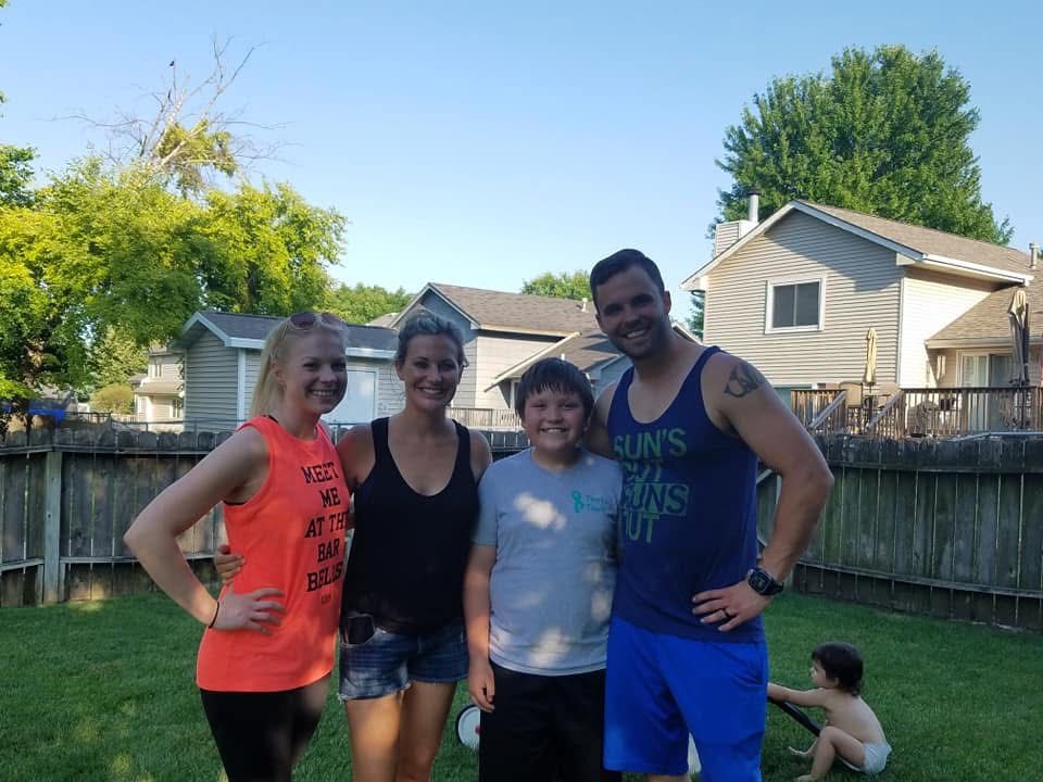 Family of four smiling together in a sunny backyard, with green grass and wooden fence in the background.
