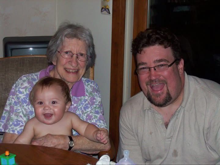 Smiling baby with elderly grandmother and bearded man posing together at a table in a cozy indoor setting.