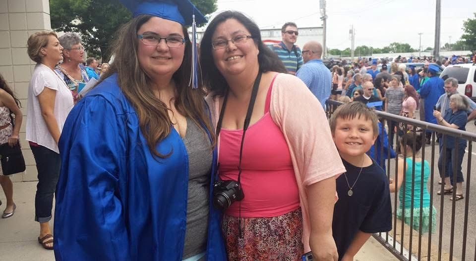 Graduate in blue cap and gown poses with two family members at an outdoor graduation ceremony.