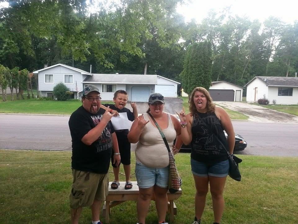 Four people posing outdoors near a residential street on a sunny summer day with green trees and houses in background.