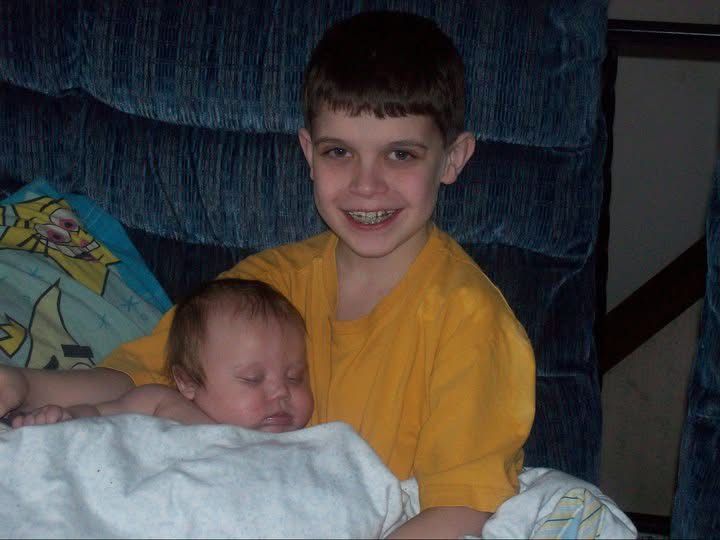 Young boy in yellow shirt smiling while holding a newborn baby wrapped in white blanket on a blue couch.