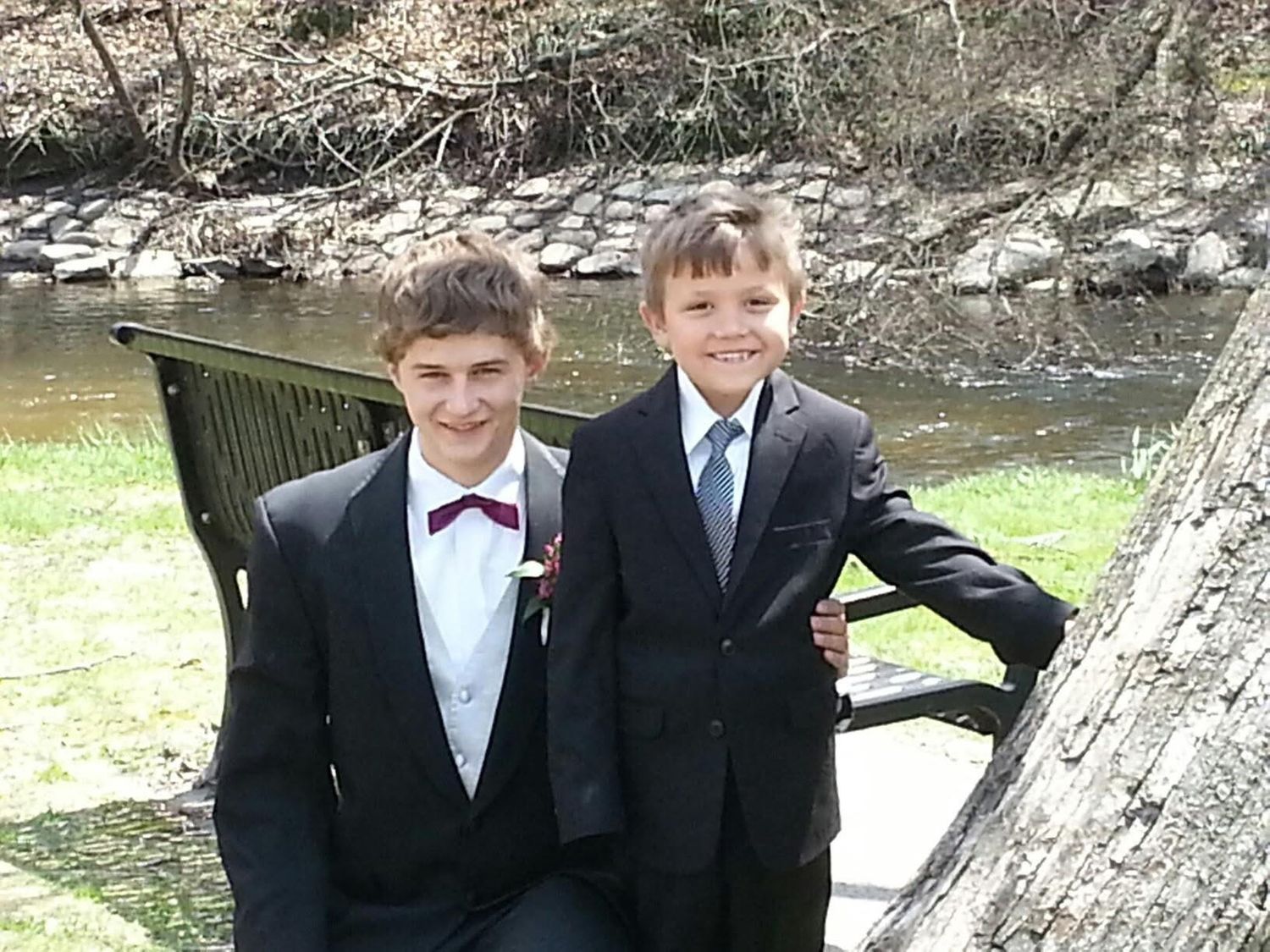 Two boys in suits and ties pose outdoors near a creek, smiling for a formal occasion photo.