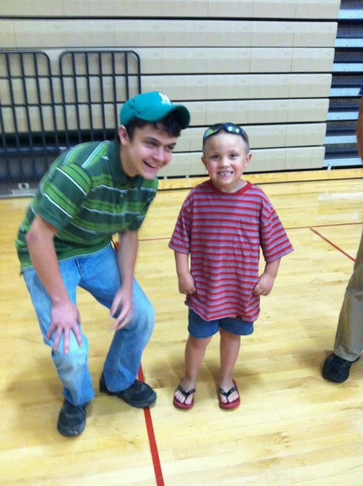 Teen boy in green striped shirt and cap poses with smiling young boy in red striped shirt on gym floor.