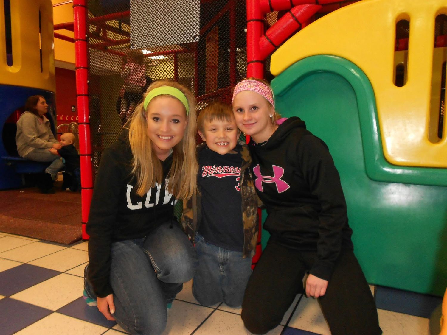 Three children smiling and posing together at a colorful indoor playground play structure.