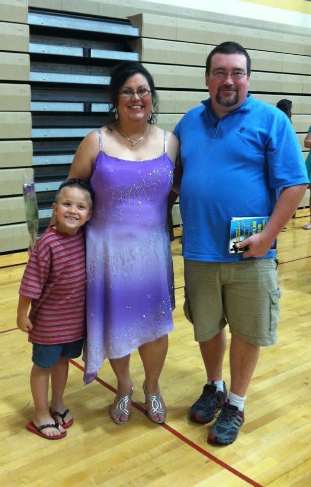 Family of three posing in a gymnasium, woman in purple dress flanked by young boy and man in blue polo shirt.