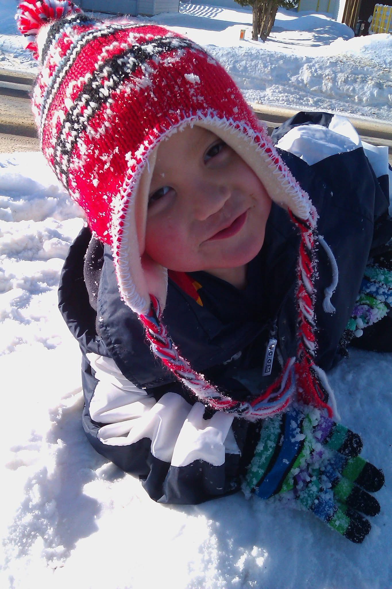 Young child in red winter hat and gloves playing in snow, wearing a dark jacket on a sunny day.