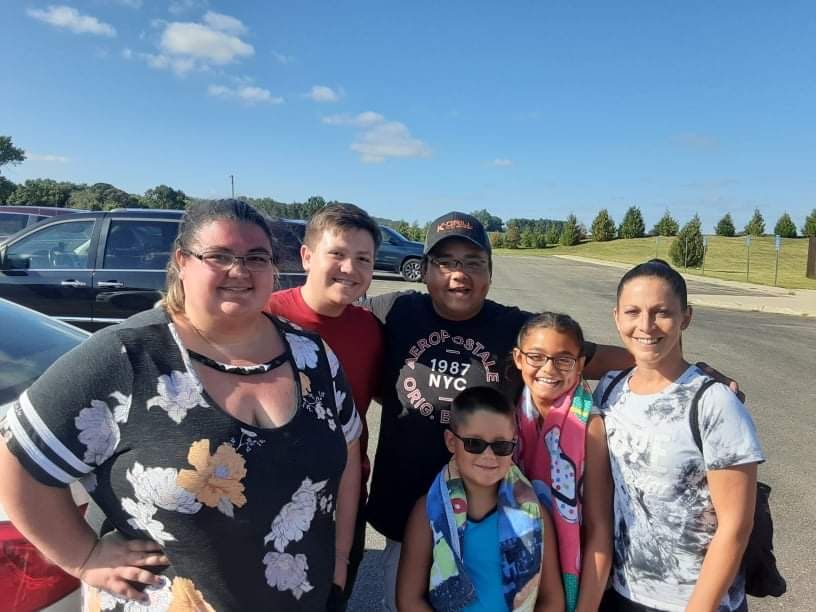 A family group of six people smiling together outdoors in a sunny parking lot near a school.
