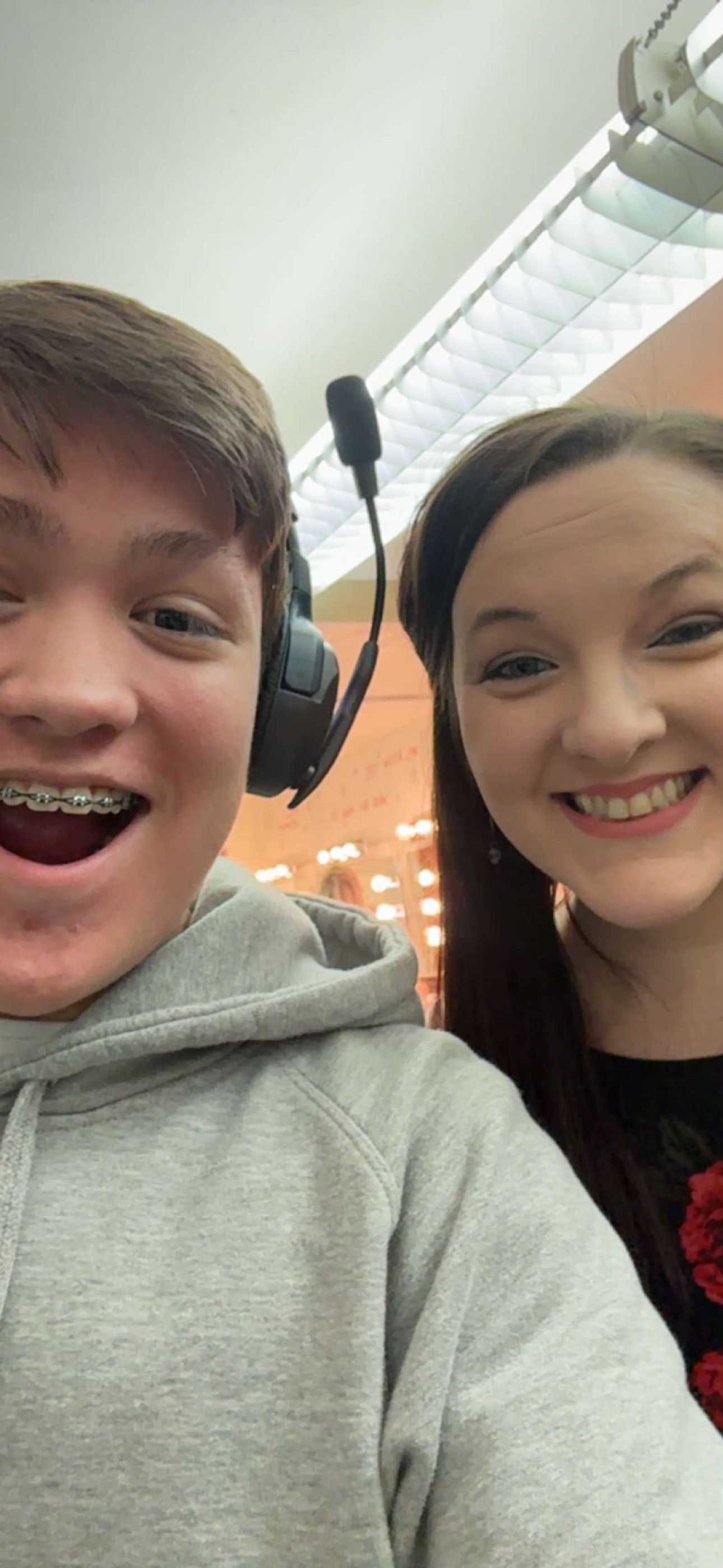 Two smiling teenagers wearing headsets take a selfie, boy in gray hoodie with braces and girl in floral top.
