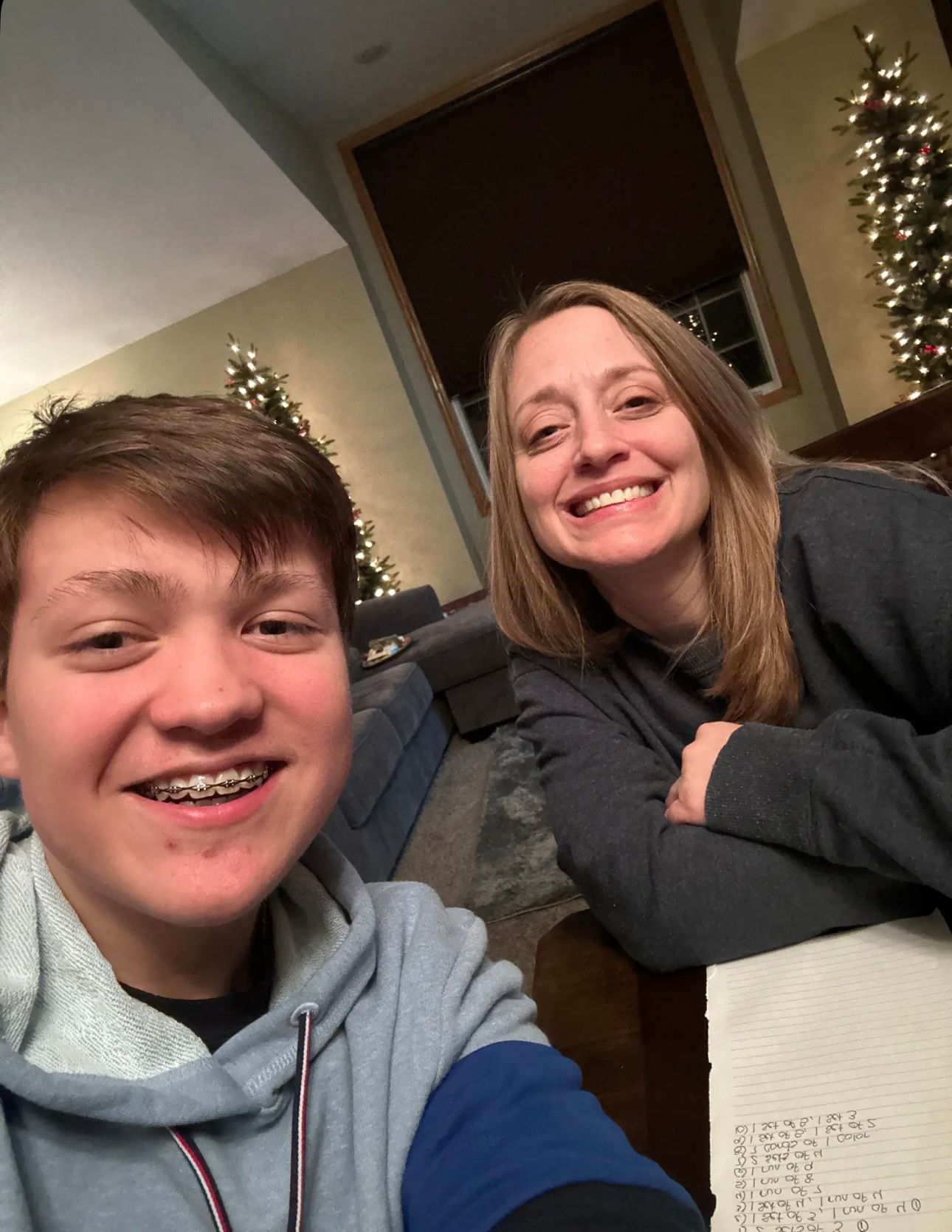 A teenage boy with braces and a woman smile together in a selfie near a decorated Christmas tree indoors.