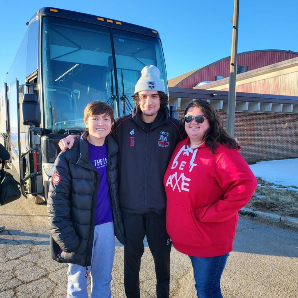 Three people posing together outdoors near a tour bus on a sunny winter day, wearing casual cold-weather clothing.