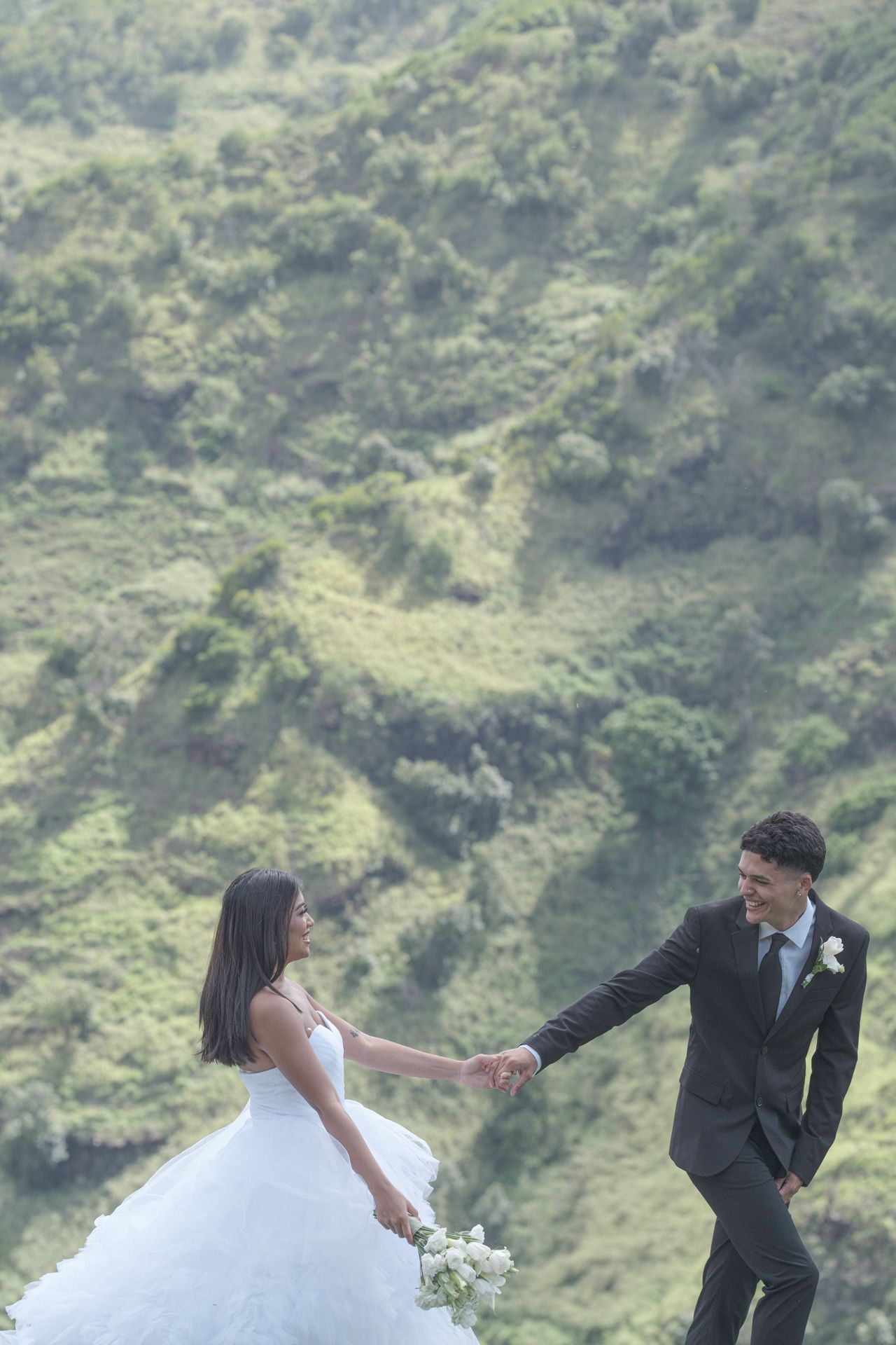 A bride and groom hold hands while posing outdoors with lush green hills in the background on their wedding day.