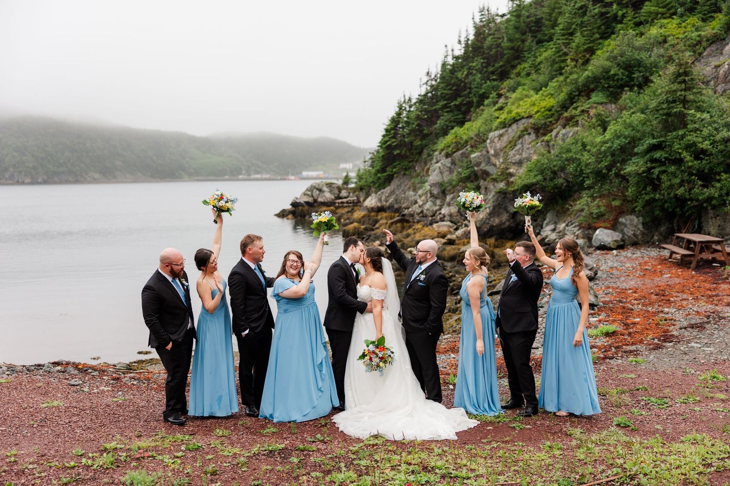 Wedding party celebrating on rocky shoreline with misty fjord backdrop, bridesmaids in blue dresses cheering.