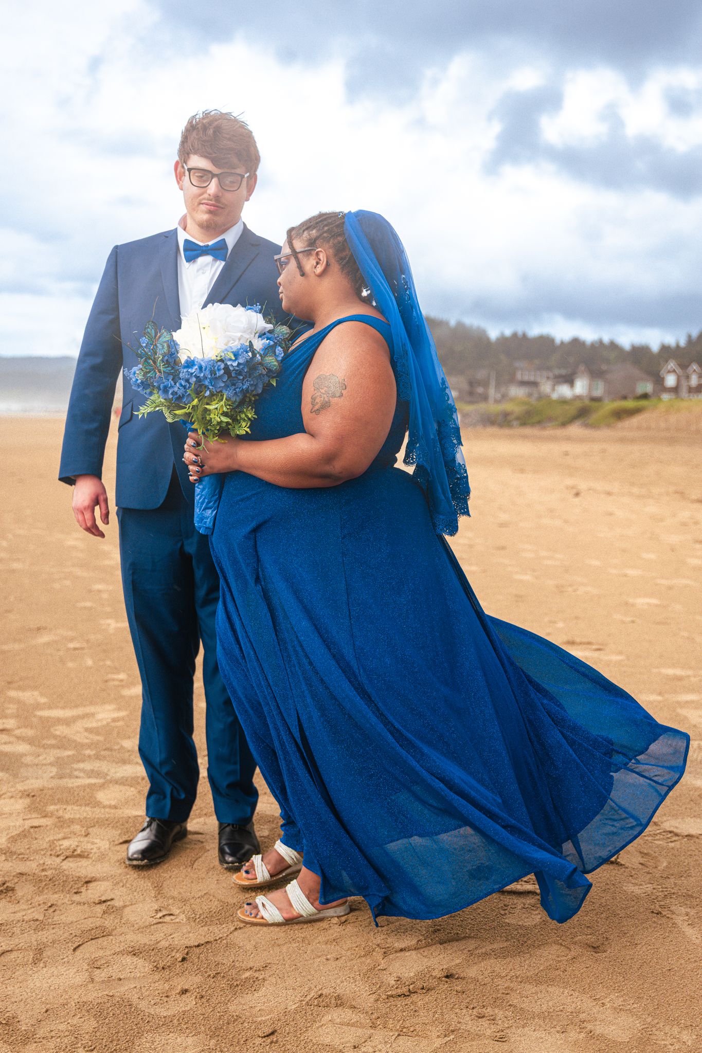 Interracial couple in matching blue wedding attire on sandy beach, bride in flowing gown with veil holding bouquet.