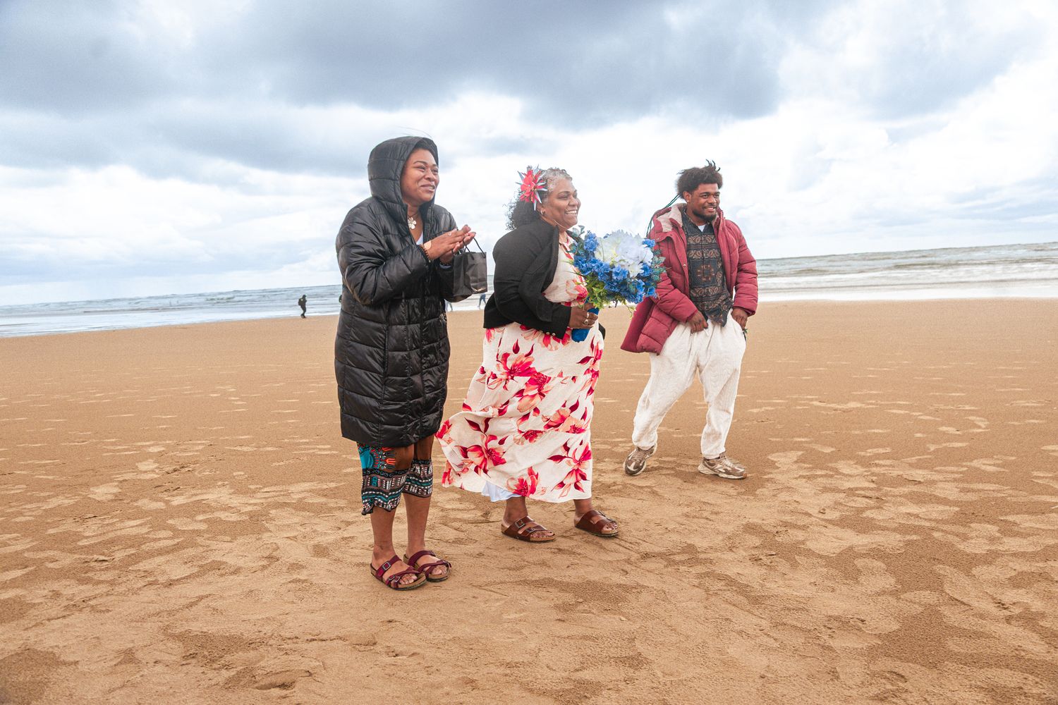Three people walking on a sandy beach on a cloudy day, dressed in colorful casual clothing.