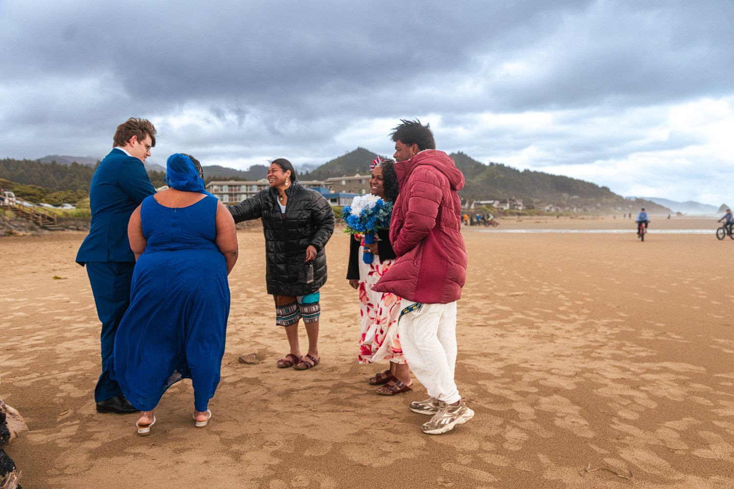 A group of people gather on a sandy beach for an outdoor wedding ceremony on an overcast day.