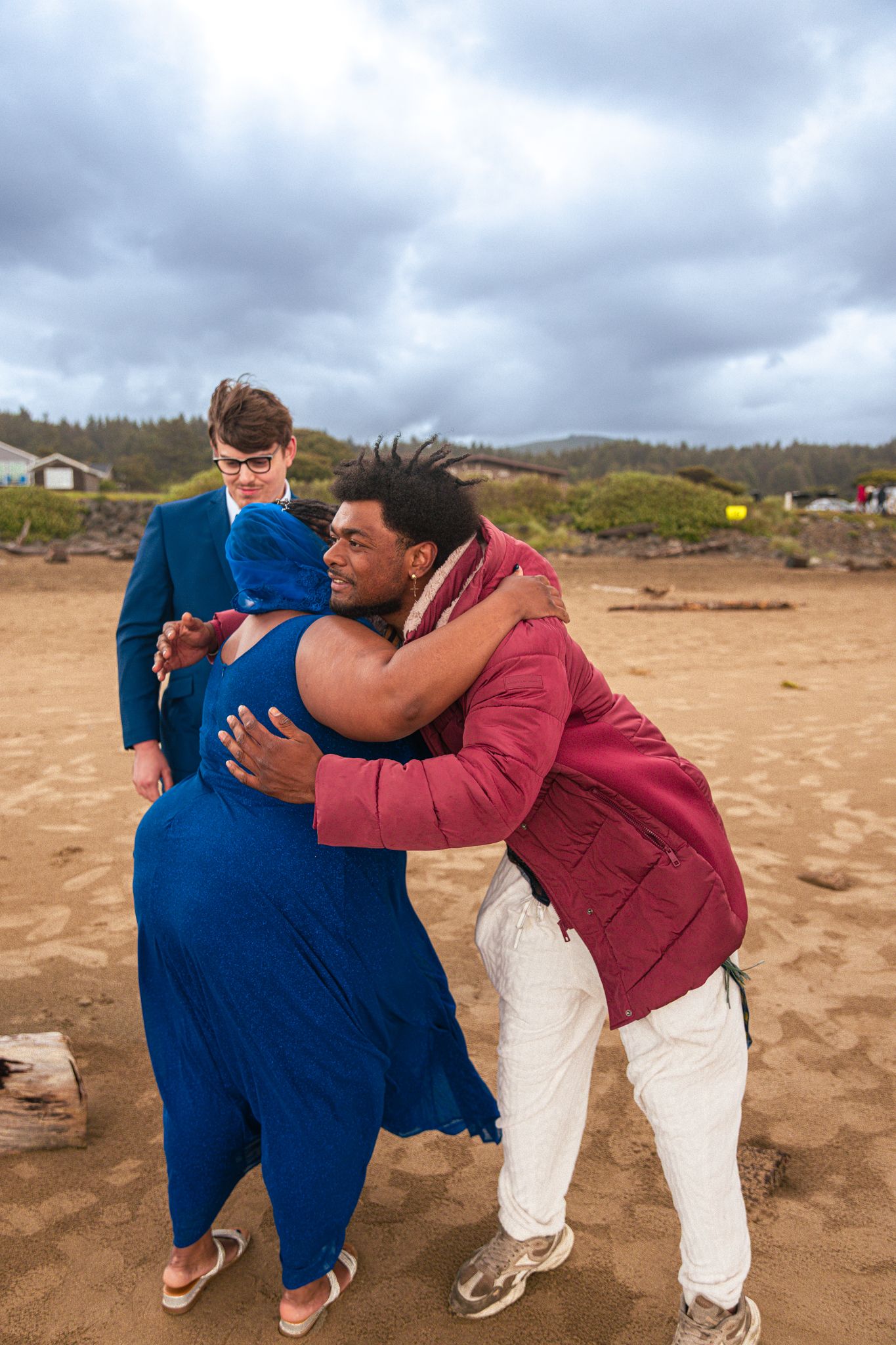 Three people sharing a warm hug on a sandy beach, dressed in blue, pink, and navy formal attire under cloudy skies.