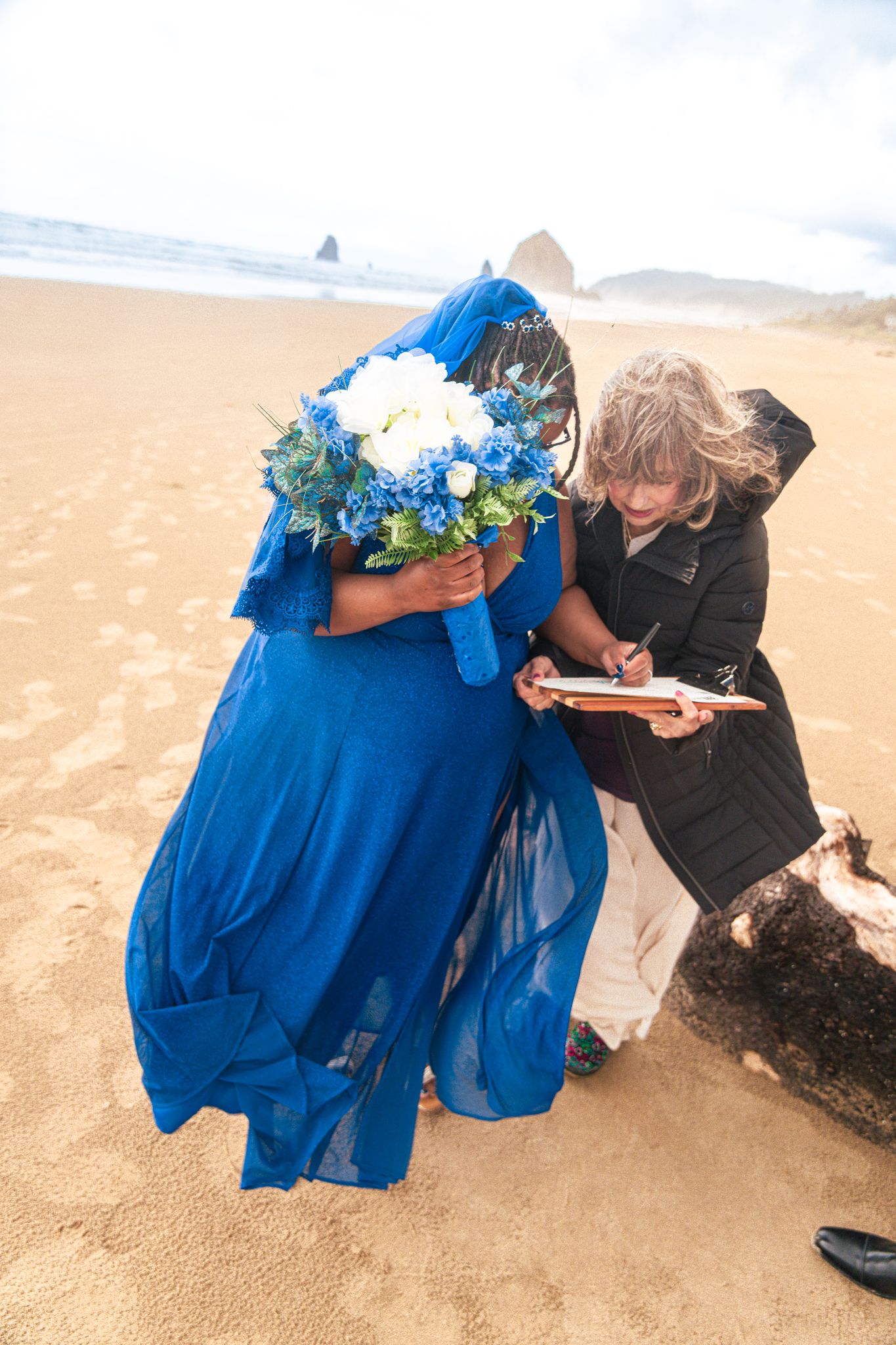 Bride in blue dress and veil signing marriage certificate on beach, holding blue and white bouquet.