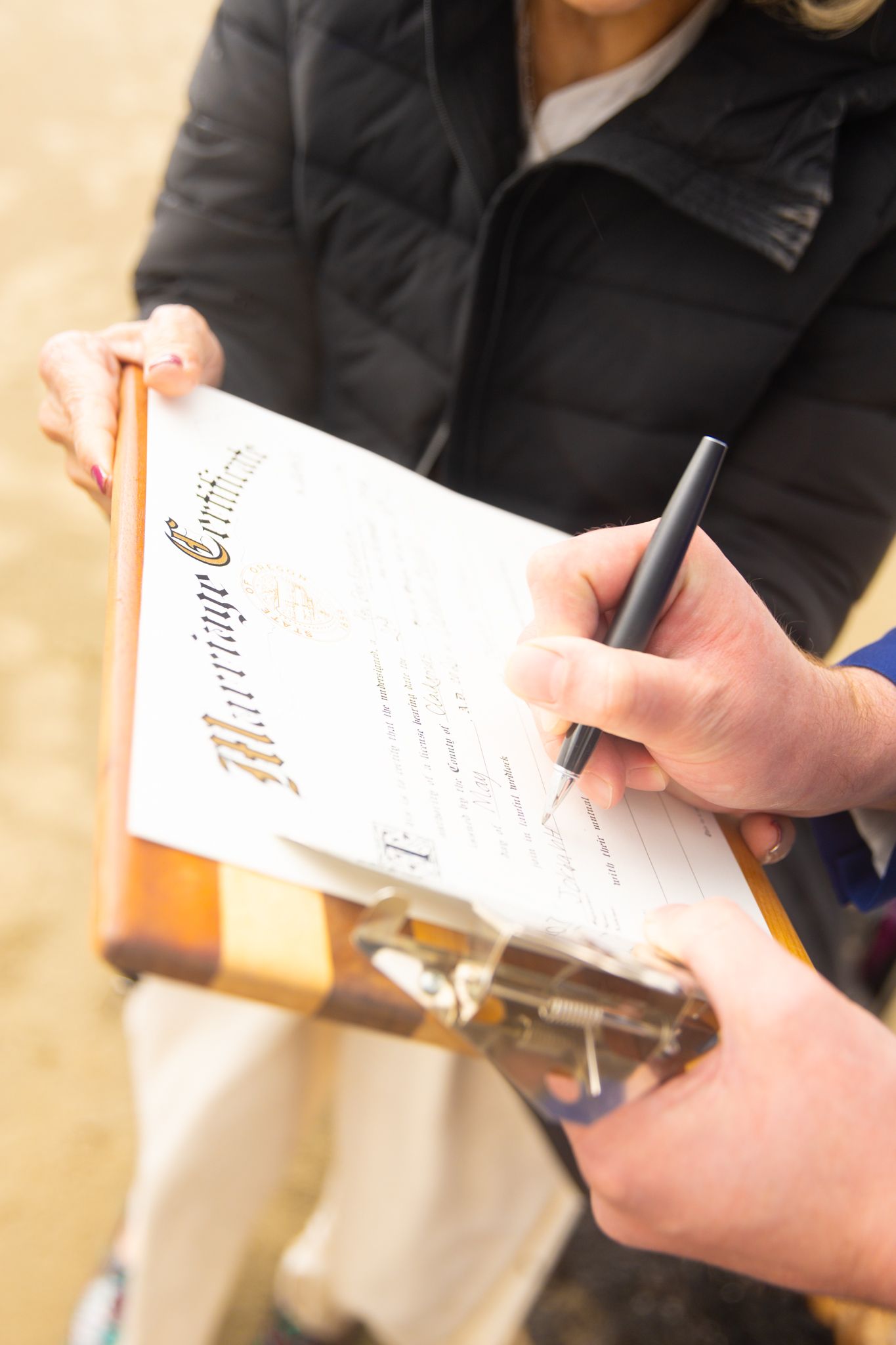 Close-up of hands signing a marriage certificate on a clipboard during a beach wedding ceremony.