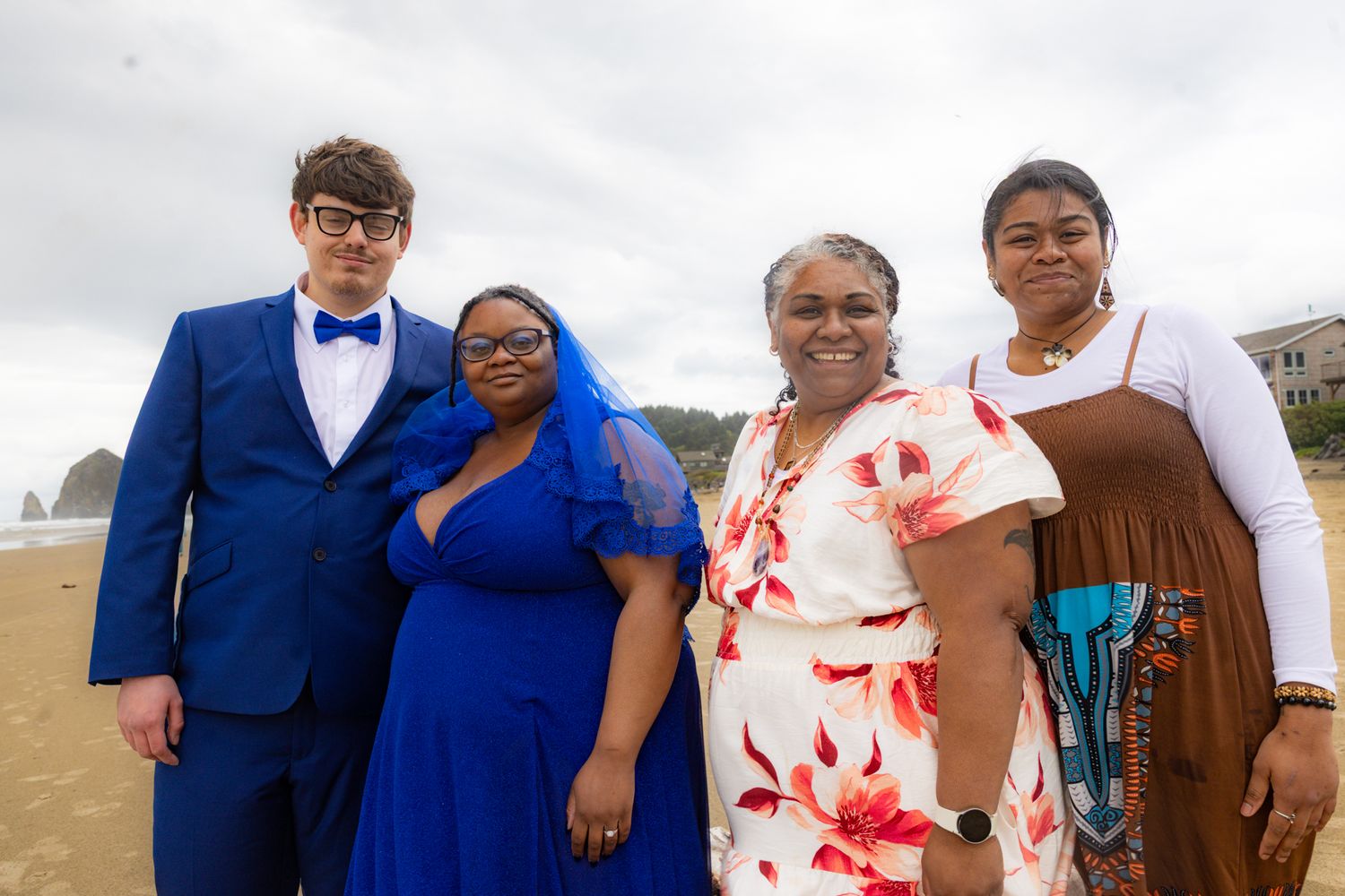 Four people posing together on a beach, dressed in formal attire including a blue suit and blue gown, on a cloudy day.