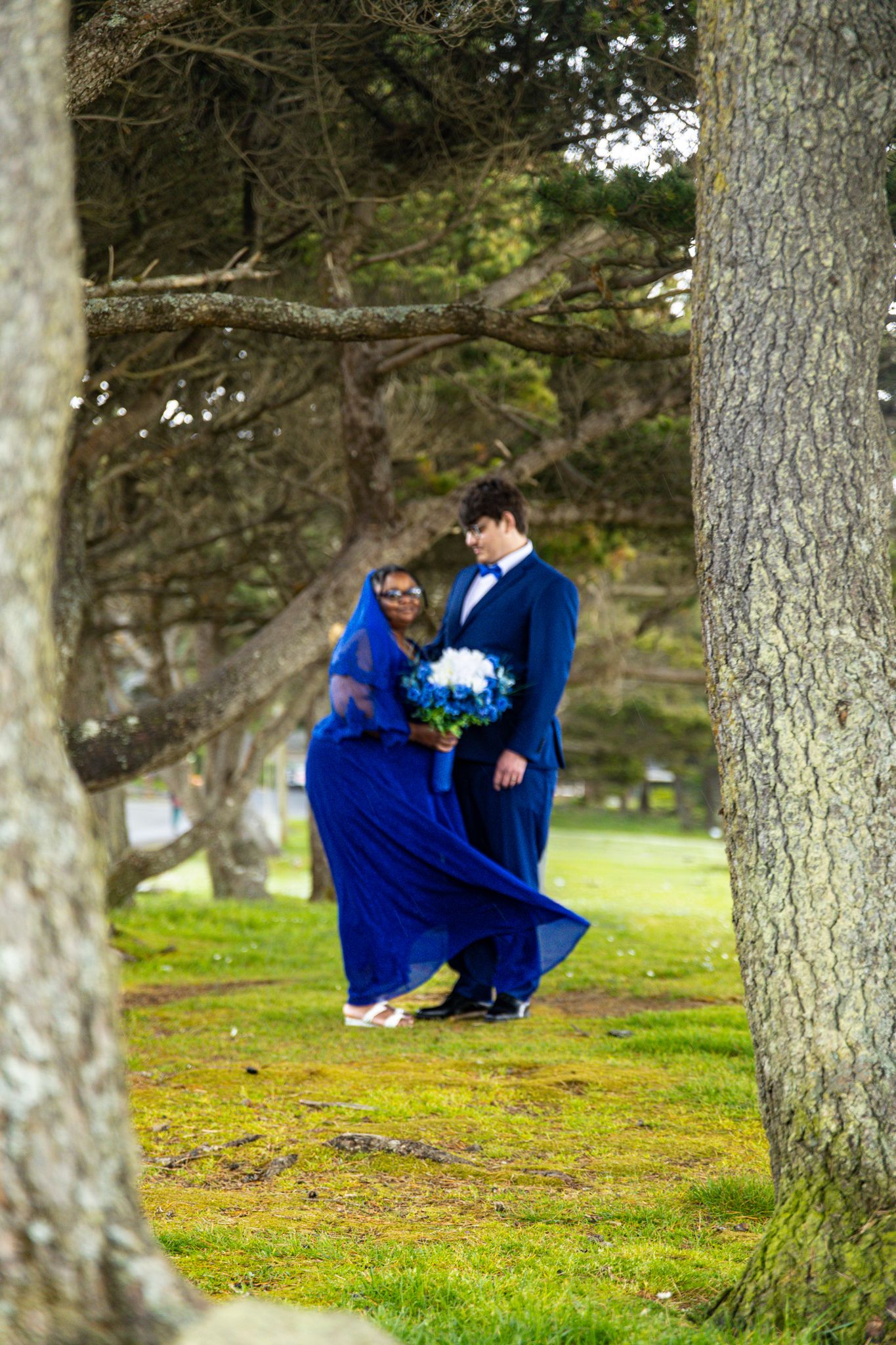 Couple in matching royal blue wedding attire embracing among trees on green grass.